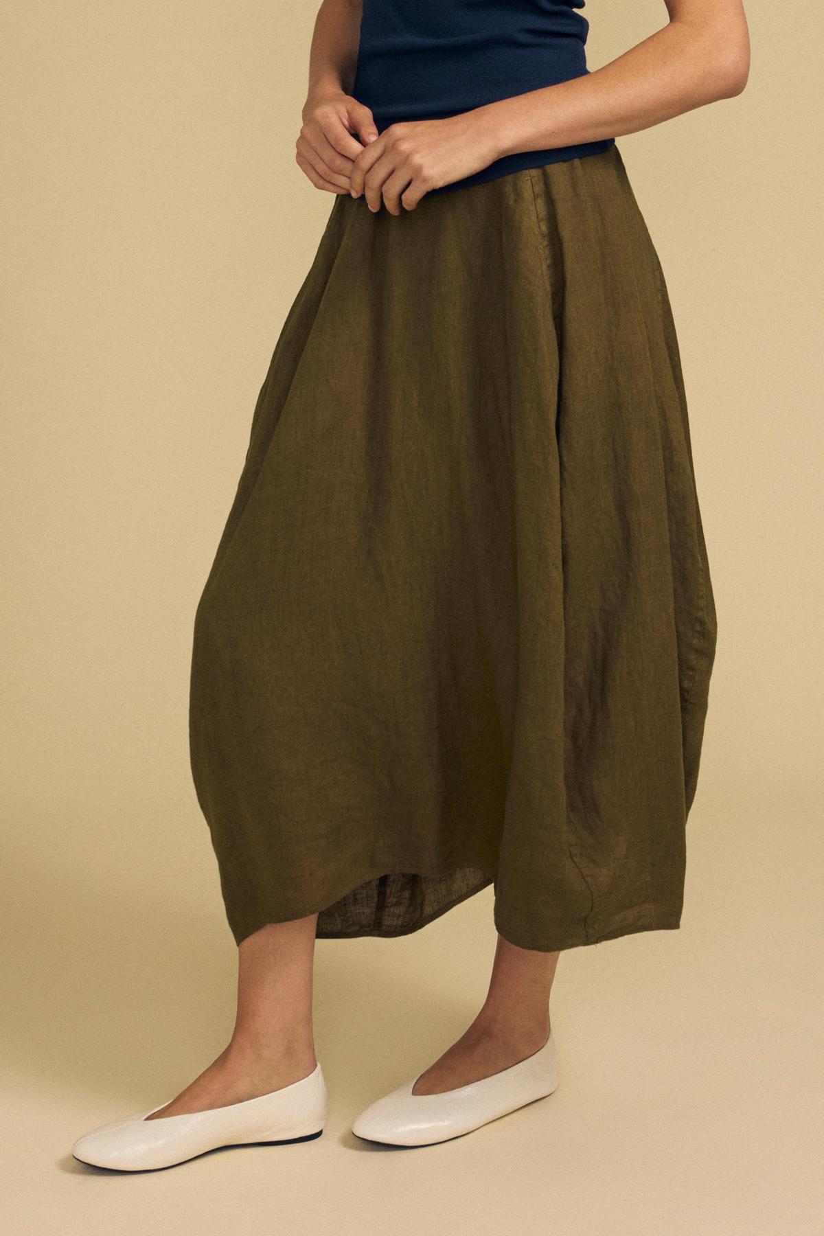 A person wearing the FAE LINEN SKIRT by Velvet by Graham & Spencer in olive green—a loose-fitting, midi-length style with an elastic waistband—paired with a navy top and white flat shoes, stands against a beige background.