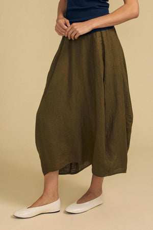 A person wearing the FAE LINEN SKIRT by Velvet by Graham & Spencer in olive green—a loose-fitting, midi-length style with an elastic waistband—paired with a navy top and white flat shoes, stands against a beige background.
