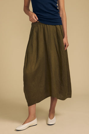 A person wearing a navy top, the FAE LINEN SKIRT by Velvet by Graham & Spencer in olive green, and white flats stands against a beige background.