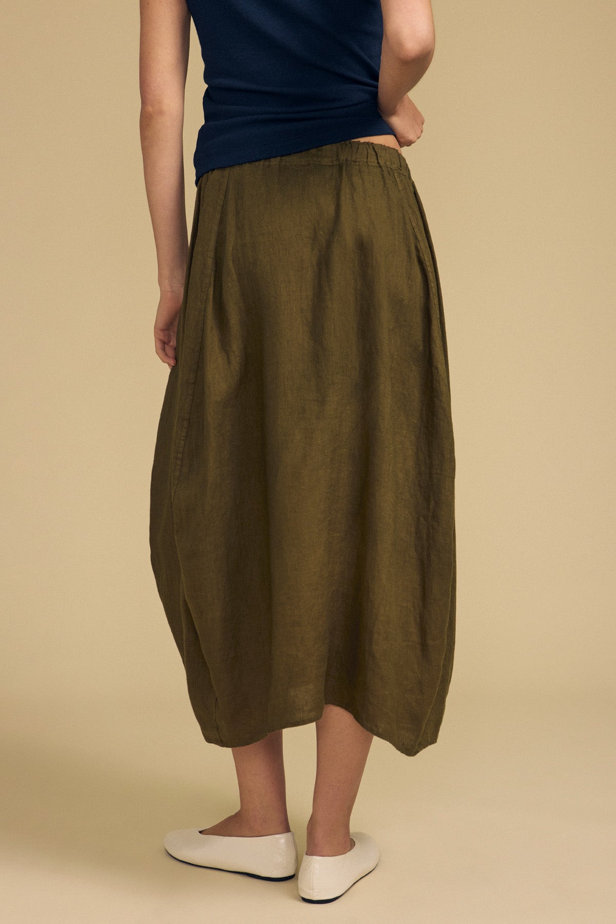 A person is shown from the back against a beige background, wearing a navy top, the Velvet by Graham & Spencer FAE LINEN SKIRT—an olive green, ankle-length A-line silhouette skirt with an elastic waistband—and white flats.