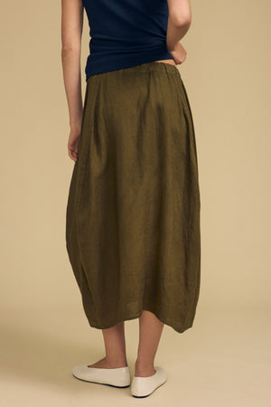 A person is shown from the back against a beige background, wearing a navy top, the Velvet by Graham & Spencer FAE LINEN SKIRT—an olive green, ankle-length A-line silhouette skirt with an elastic waistband—and white flats.