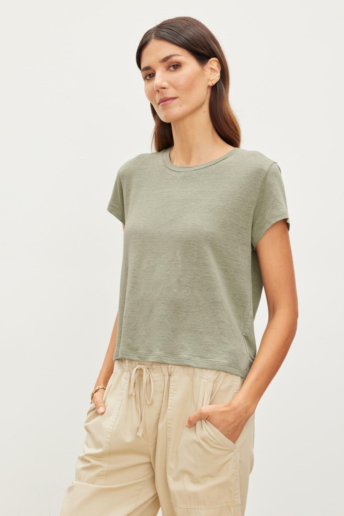   A woman in a Velvet by Graham & Spencer CASEY LINEN KNIT CREW NECK TEE and beige drawstring pants stands with her hands in her pockets, looking at the camera with a subtle expression. 