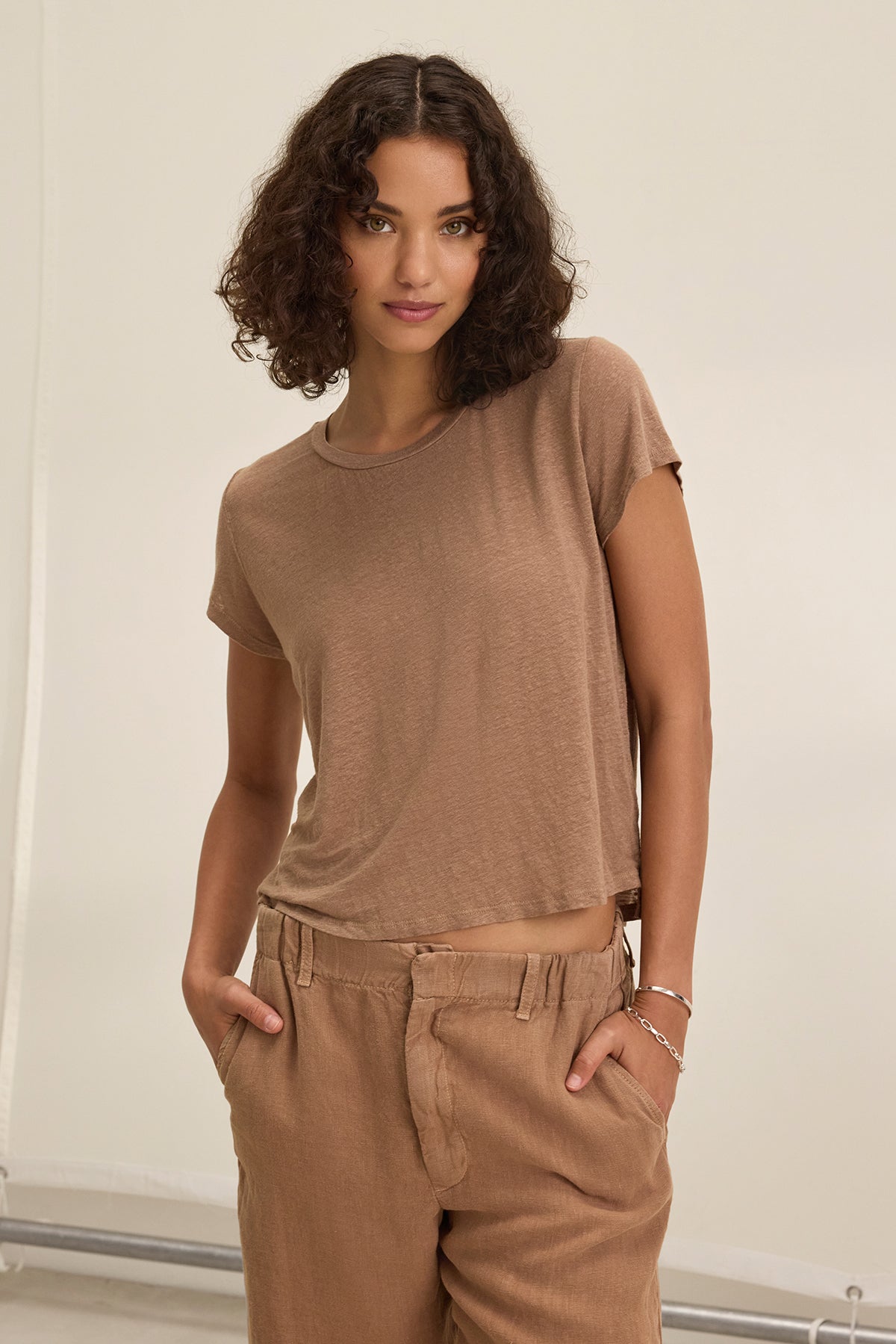 A person with curly hair wears the CASEY LINEN KNIT TEE by Velvet by Graham & Spencer and matching pants, both providing a flawless fit, standing with hands in pockets against a neutral background.-38662914638017