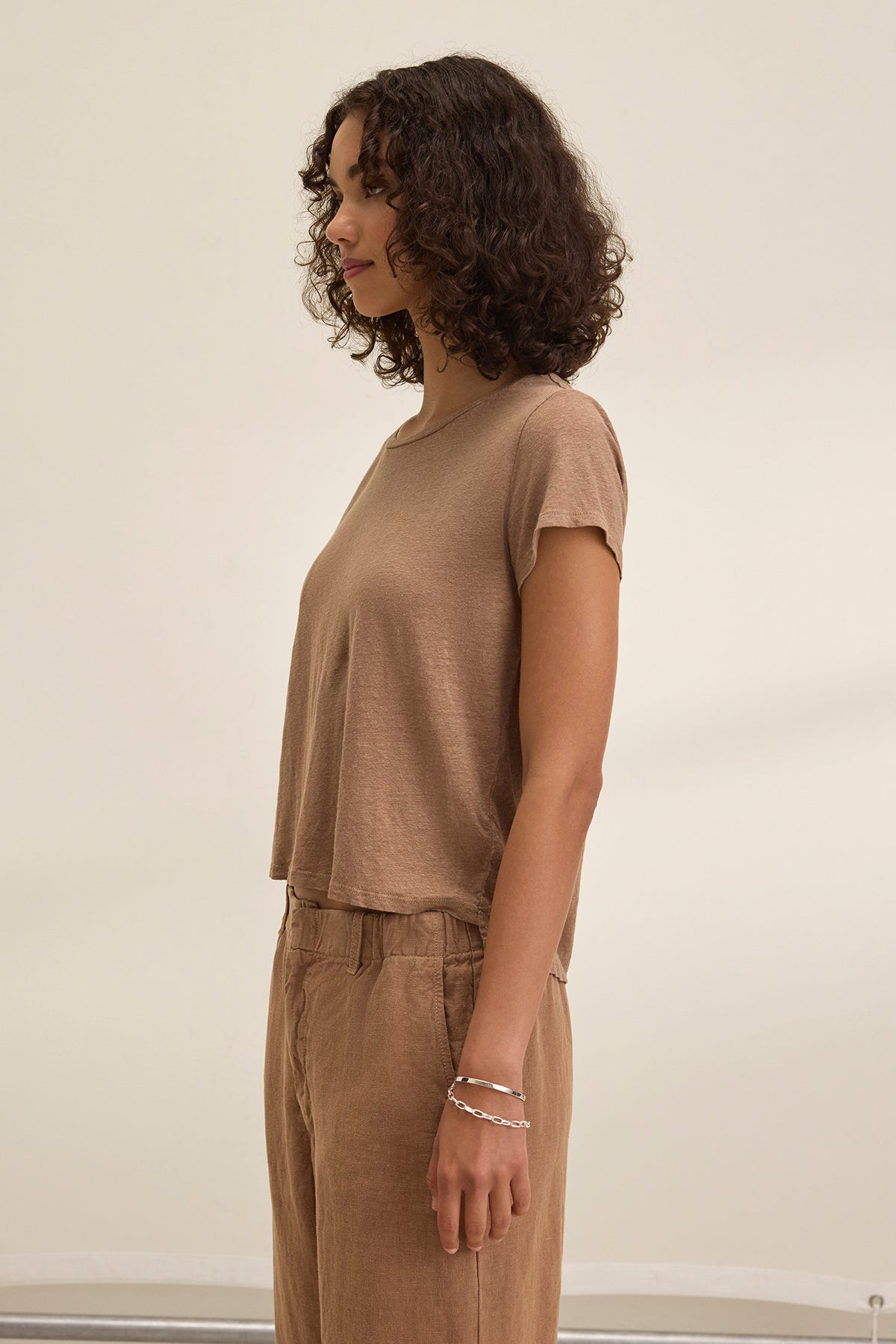 A person with curly hair stands in profile, wearing the CASEY LINEN KNIT TEE by Velvet by Graham & Spencer and matching pants in brown, exuding effortless style against a plain background.-38662914703553