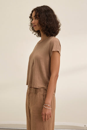 A person with curly hair stands in profile, wearing the CASEY LINEN KNIT TEE by Velvet by Graham & Spencer and matching pants in brown, exuding effortless style against a plain background.