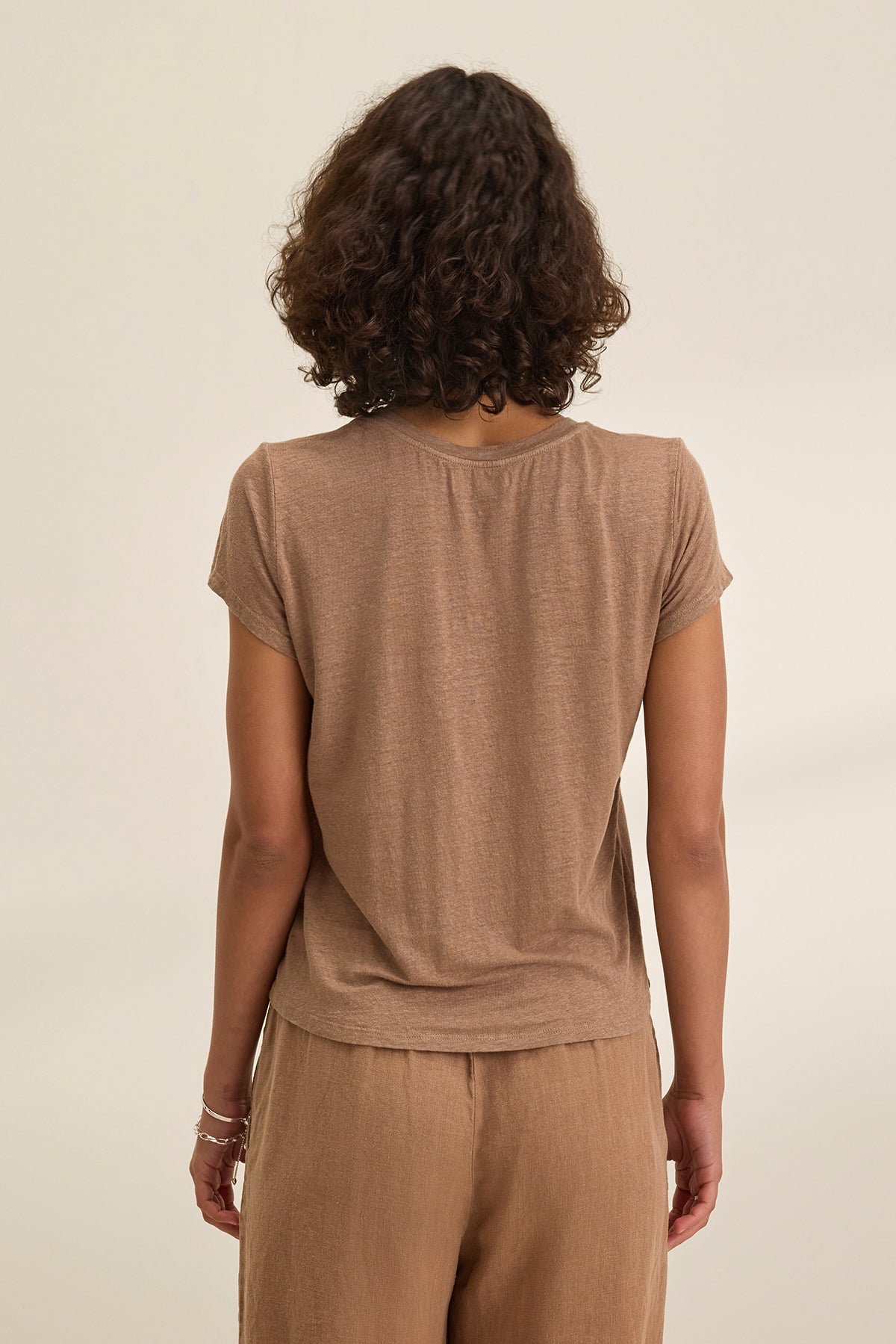 Curly-haired individual in a Velvet by Graham & Spencer CASEY LINEN KNIT TEE and perfectly fitting pants stands back to the camera against a plain background.-38662914736321