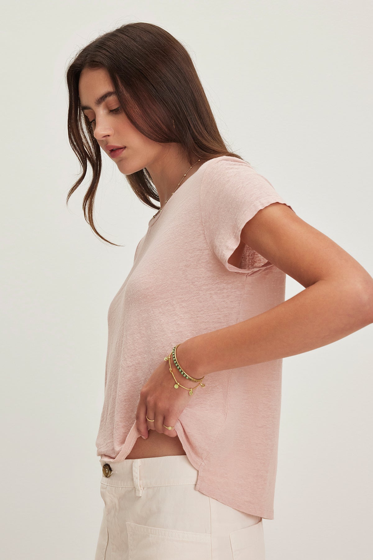 Woman with long brown hair in a CASEY LINEN KNIT TEE by Velvet by Graham & Spencer and white pants stands against a plain background, one hand on her hip, showcasing effortless style.-38662914801857