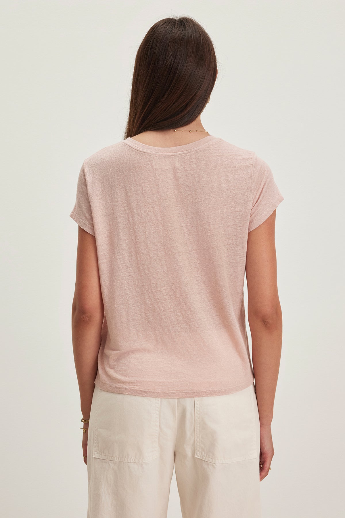 Person with long brown hair wears the CASEY LINEN KNIT TEE by Velvet by Graham & Spencer in light pink with a high-low hemline, paired with white pants, shown from the back against a plain background.-38662914834625
