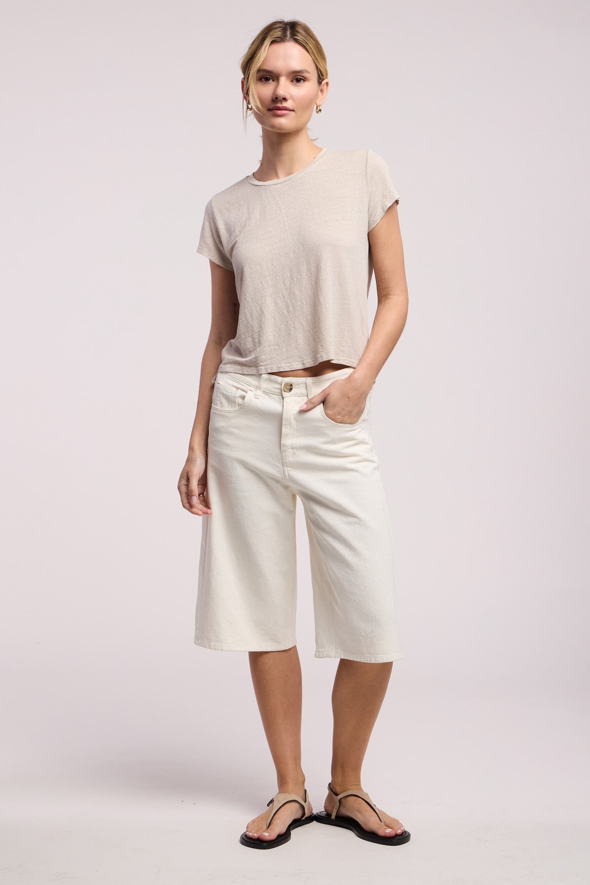 A woman stands against a plain background wearing the CASEY TEE by Velvet by Graham & Spencer in beige, paired with knee-length white shorts and black sandals, with one hand in her pocket.-41371566637249