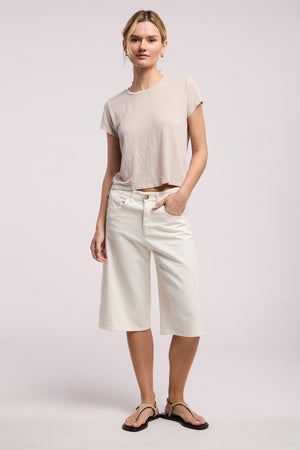 A woman stands against a plain background wearing the CASEY TEE by Velvet by Graham & Spencer in beige, paired with knee-length white shorts and black sandals, with one hand in her pocket.