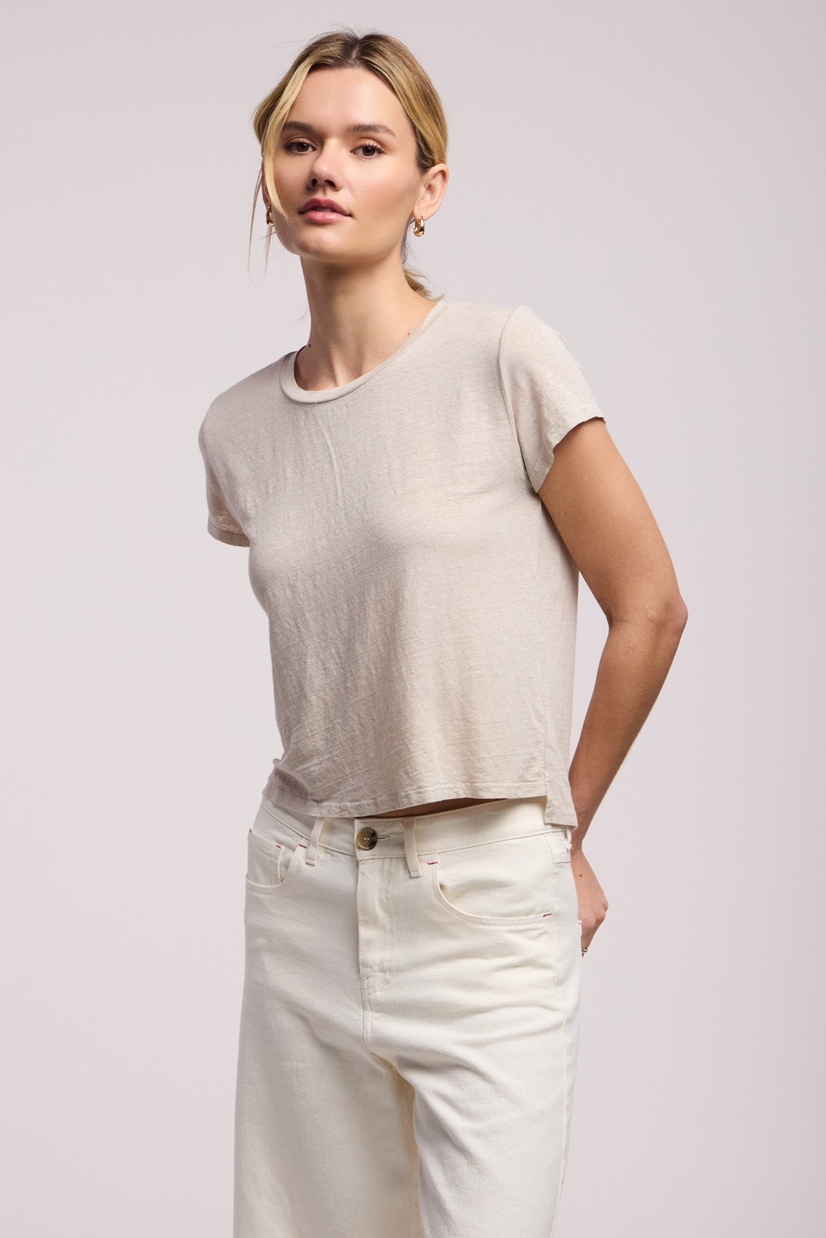 A person faces forward against a plain background, wearing the Velvet by Graham & Spencer CASEY TEE in light beige linen with a classic high-low hemline, paired with high-waisted off-white jeans.-41371566538945