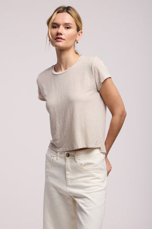 A person faces forward against a plain background, wearing the Velvet by Graham & Spencer CASEY TEE in light beige linen with a classic high-low hemline, paired with high-waisted off-white jeans.