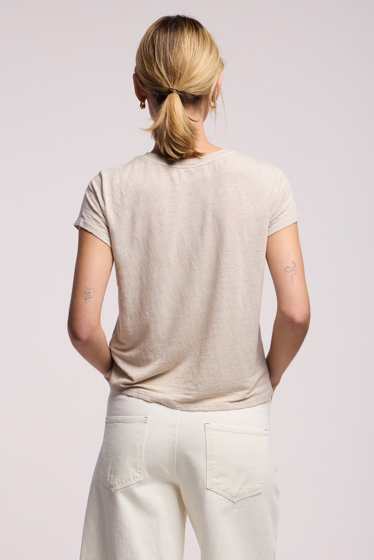 A person with blonde hair in a ponytail, wearing the CASEY TEE by Velvet by Graham & Spencer—a light beige linen knit top with a high-low hem—and white pants stands facing away. Two small tattoos are visible on their upper arms.