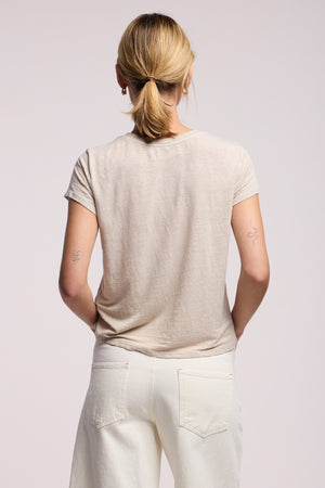 A person with blonde hair in a ponytail, wearing the CASEY TEE by Velvet by Graham & Spencer—a light beige linen knit top with a high-low hem—and white pants stands facing away. Two small tattoos are visible on their upper arms.