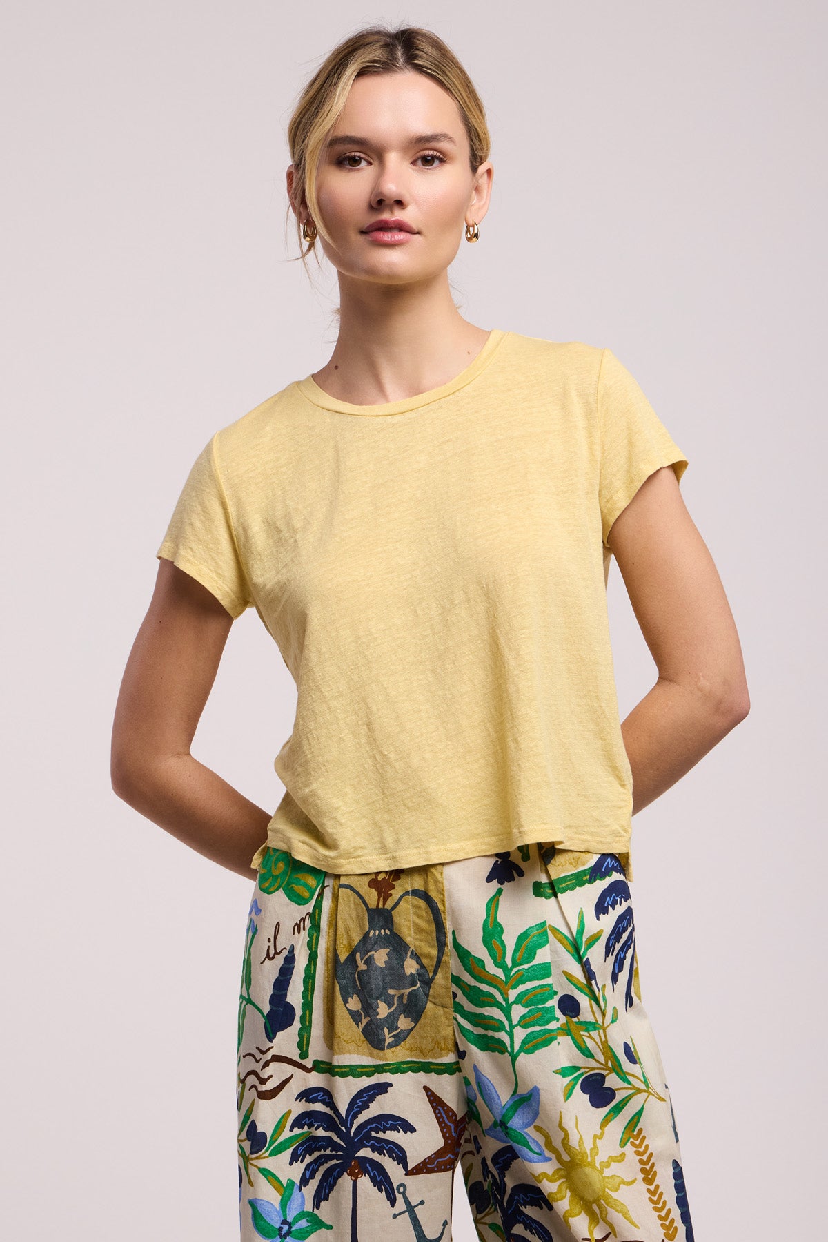 A woman faces forward, wearing the CASEY TEE by Velvet by Graham & Spencer—a light yellow linen knit with a classic high-low hem—and high-waisted pants with a colorful botanical print.
