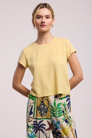 A woman faces forward, wearing the CASEY TEE by Velvet by Graham & Spencer—a light yellow linen knit with a classic high-low hem—and high-waisted pants with a colorful botanical print.
