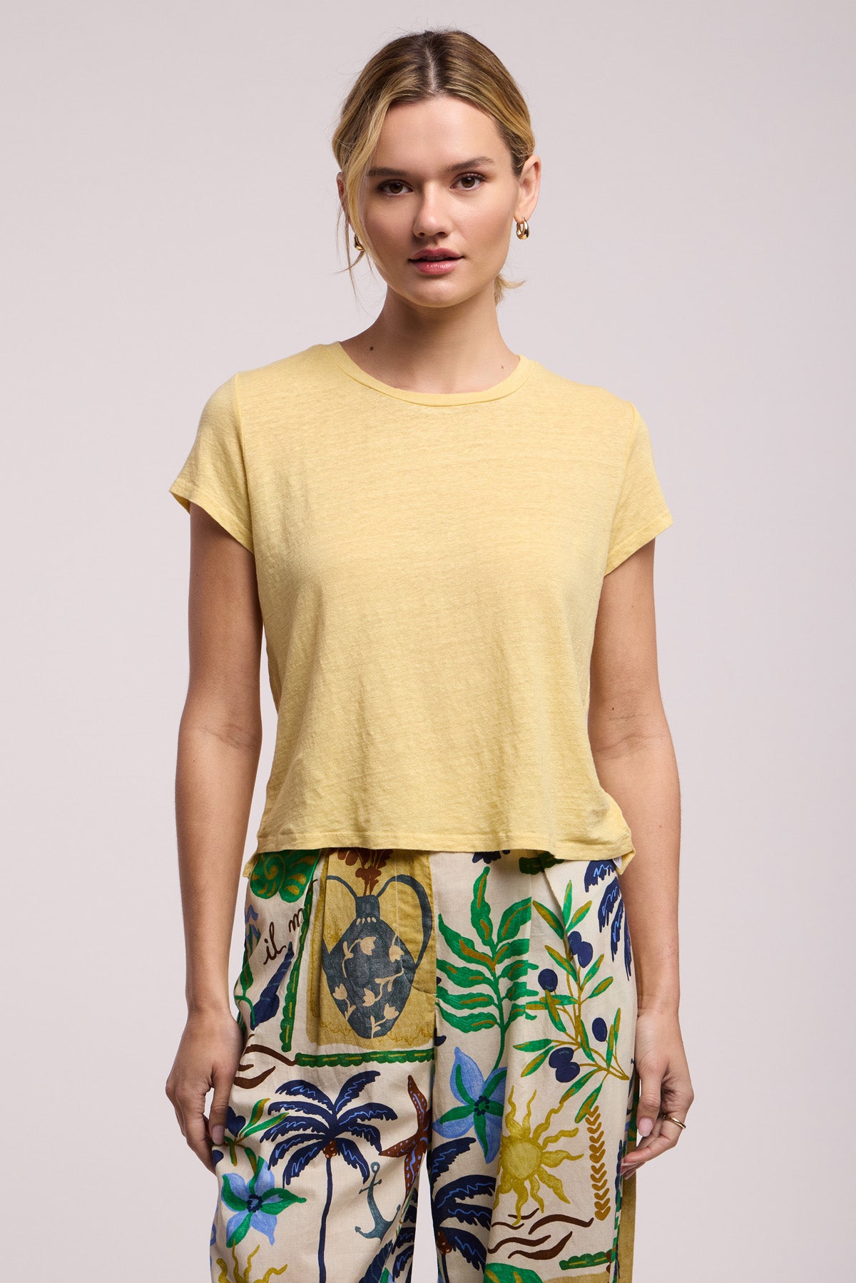 A person stands facing forward, wearing the light yellow CASEY TEE by Velvet by Graham & Spencer and high-waisted pants with plant and vase patterns, set against a plain background.