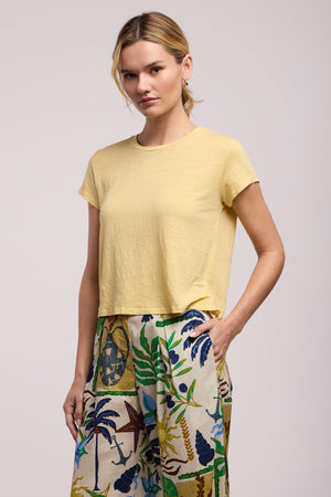 Wearing the CASEY TEE by Velvet by Graham & Spencer in yellow and high-waisted, wide-leg pants with a vibrant tropical print, this person stands confidently with one hand in their pocket.