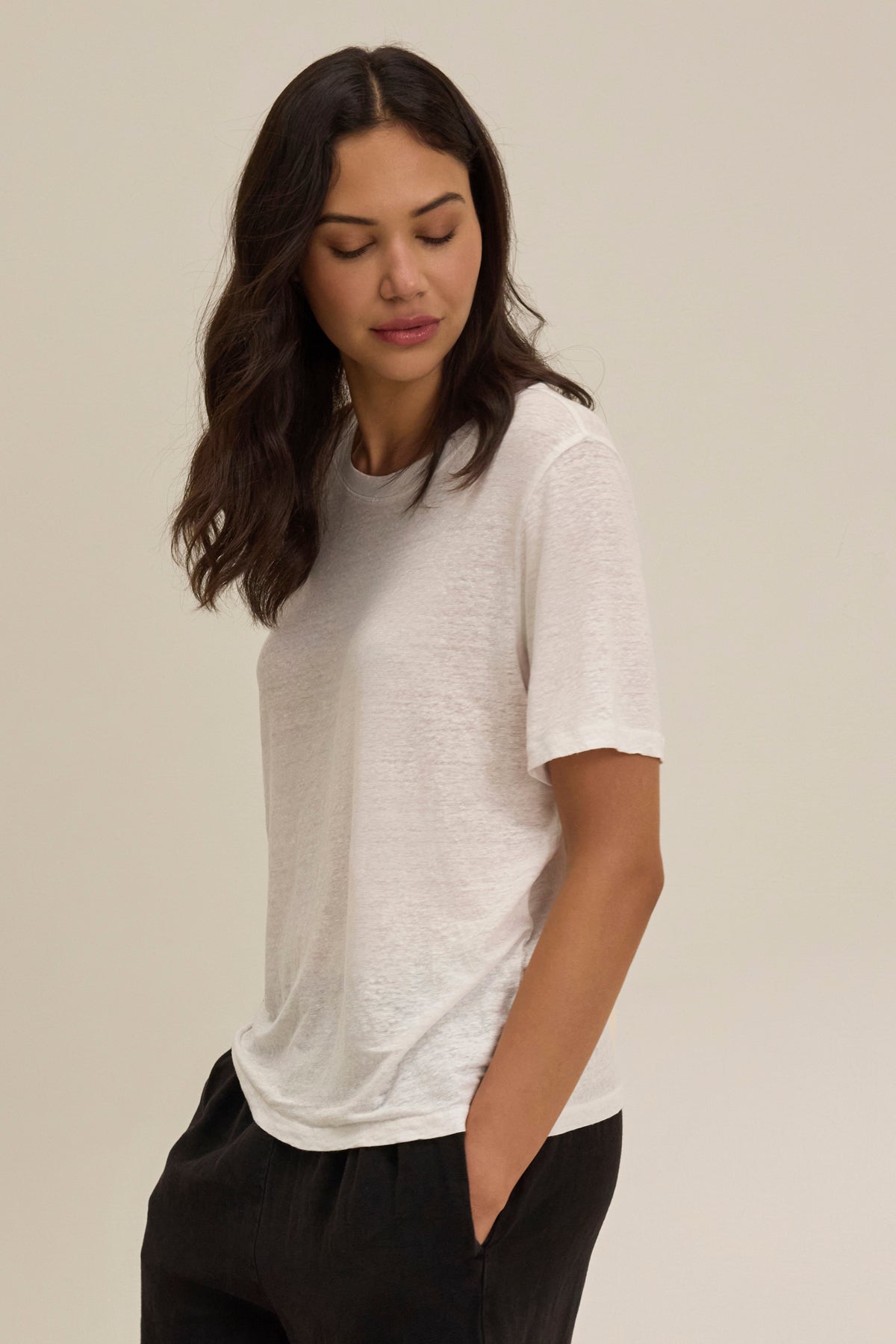 A person with long, dark hair in a HERITAGE LINEN KNIT TEE by Velvet by Graham & Spencer and relaxed fit black pants stands against a plain background, looking down with a neutral expression.-38879369199809