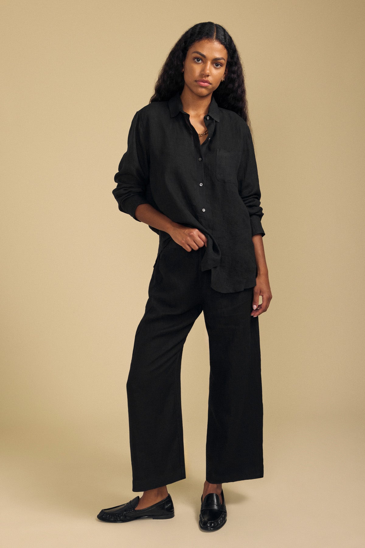 A person stands against a plain beige background wearing a loose black button-up shirt, black slip-on shoes, and the LOLA LINEN PANT by Velvet by Graham & Spencer—wide-leg linen pants featuring an elastic waist.