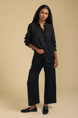 A person stands against a plain beige background wearing a loose black button-up shirt, black slip-on shoes, and the LOLA LINEN PANT by Velvet by Graham & Spencer—wide-leg linen pants featuring an elastic waist.