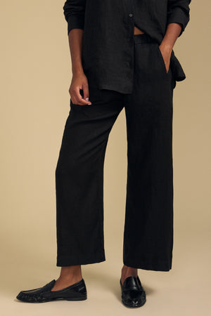 A person stands against a beige background wearing a loose-fitting black shirt, black loafers, and the LOLA LINEN PANT by Velvet by Graham & Spencer—black wide-leg pants with an elastic waist—one hand casually placed in their pocket.