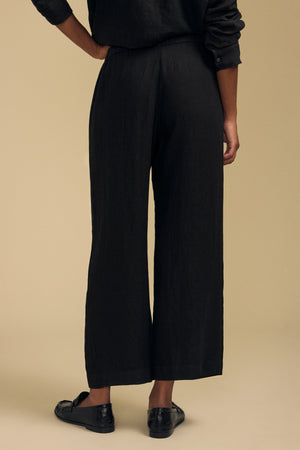 Wearing the Velvet by Graham & Spencer LOLA LINEN PANT in black—with a wide-leg silhouette and elastic waist—a person stands facing away, paired with a matching black long-sleeve top and black loafers against a beige background.