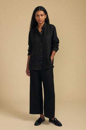 A person stands against a beige background wearing a black button-up shirt and the LOLA LINEN PANT by Velvet by Graham & Spencer—a pair of wide-leg, elastic-waist linen pants in black—paired with black shoes. Their hands are relaxed at their sides and their expression is neutral.