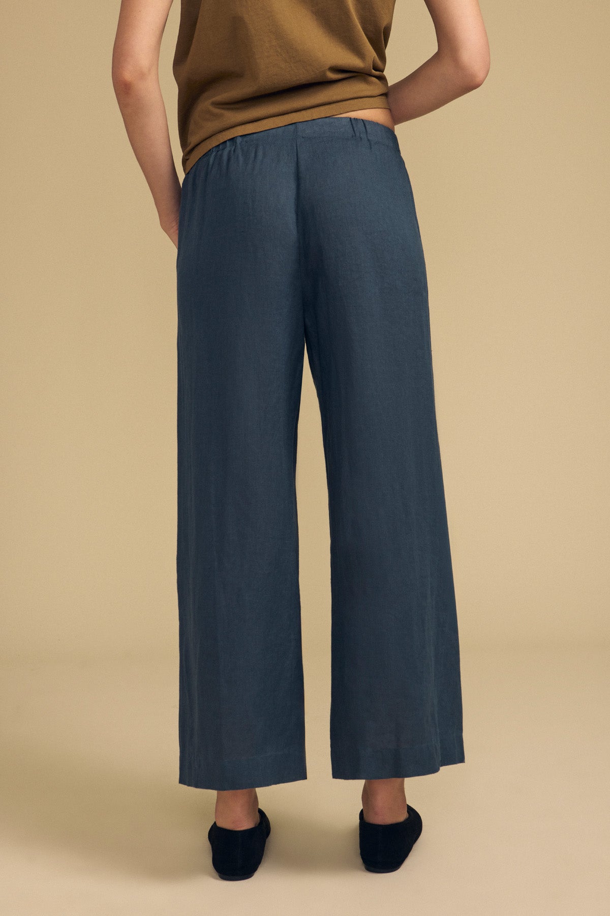 A person stands with their back to the camera, wearing a brown top and the LOLA LINEN PANT by Velvet by Graham & Spencer—loose, blue linen pants featuring an elastic waist and wide legs—paired with black shoes, set against a plain beige background.-41964619923649