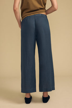 A person stands with their back to the camera, wearing a brown top and the LOLA LINEN PANT by Velvet by Graham & Spencer—loose, blue linen pants featuring an elastic waist and wide legs—paired with black shoes, set against a plain beige background.