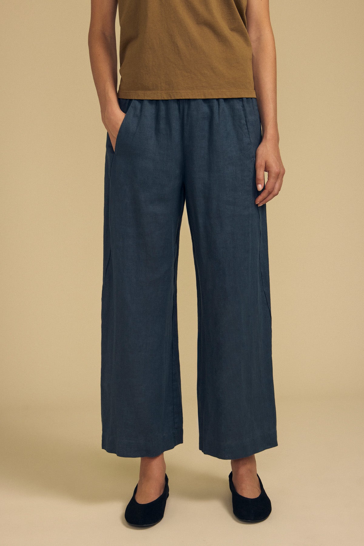 A person stands with hands in pockets, wearing a brown shirt, the Velvet by Graham & Spencer LOLA LINEN PANT in blue—a loose, wide-leg style with an elastic waist—paired with black shoes against a beige background.-41964619989185