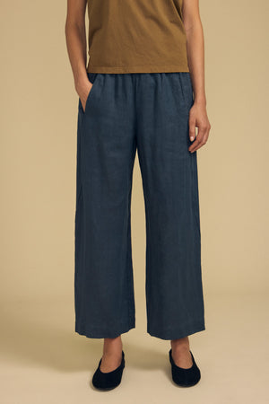 A person stands with hands in pockets, wearing a brown shirt, the Velvet by Graham & Spencer LOLA LINEN PANT in blue—a loose, wide-leg style with an elastic waist—paired with black shoes against a beige background.