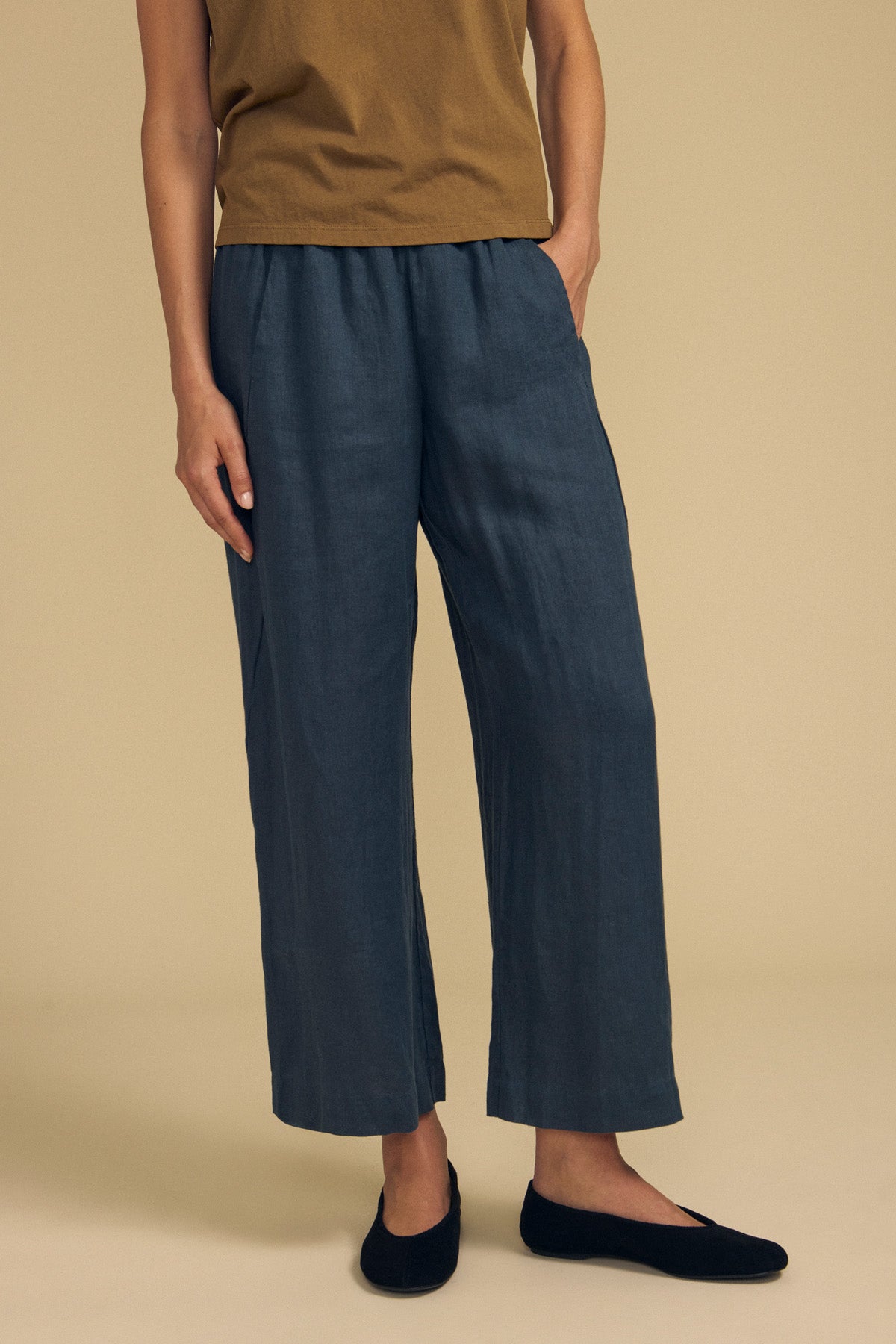 Wearing the LOLA LINEN PANT by Velvet by Graham & Spencer in a loose-fitting, dark blue style with an elastic waist and wide leg, paired with a brown short-sleeve top and black flats, the person stands against a plain beige background. The head and lower arms are out of frame.-41964620054721