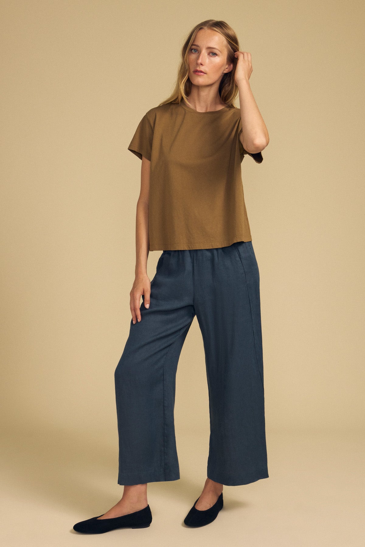A woman stands against a beige background wearing a brown short-sleeve top, black flats, and the LOLA LINEN PANT by Velvet by Graham & Spencer—loose blue-gray linen pants featuring an elastic waist and wide leg—with one hand touching her hair.