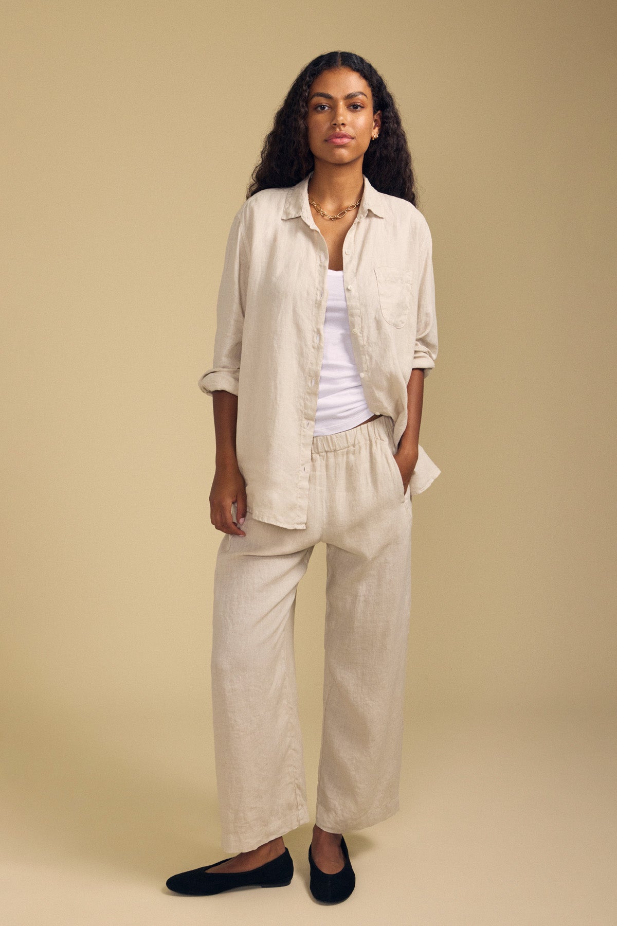 A person stands against a beige background, wearing a light-colored button-up shirt over a white top, the LOLA LINEN PANT by Velvet by Graham & Spencer—relaxed wide leg linen pants with an elastic waist—and black shoes.-41959882653889