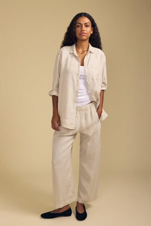 A person stands against a beige background, wearing a light-colored button-up shirt over a white top, the LOLA LINEN PANT by Velvet by Graham & Spencer—relaxed wide leg linen pants with an elastic waist—and black shoes.