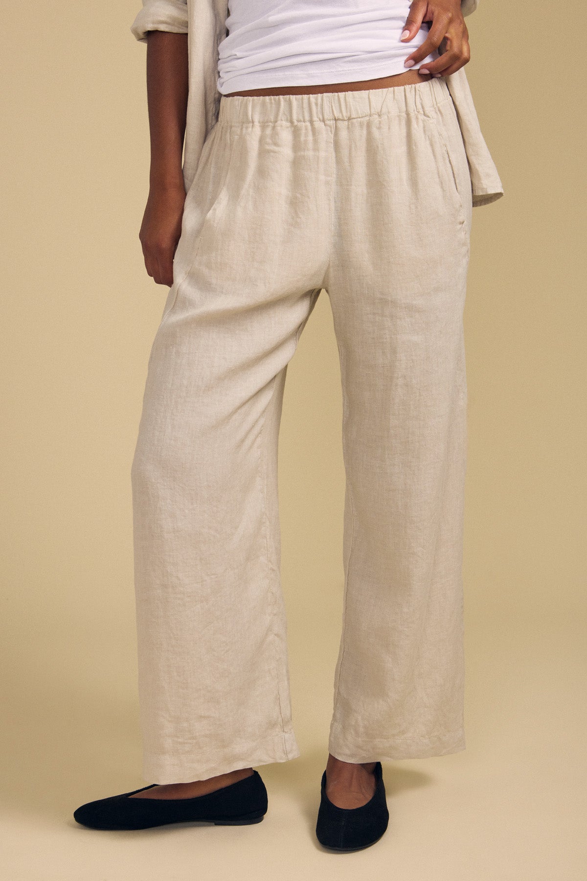 A person stands against a plain beige background wearing the Velvet by Graham & Spencer LOLA LINEN PANT in a loose, light beige wide-leg style, paired with a white top and black flats. Their partially visible hands emphasize the relaxed fit of the pants.-41959882621121