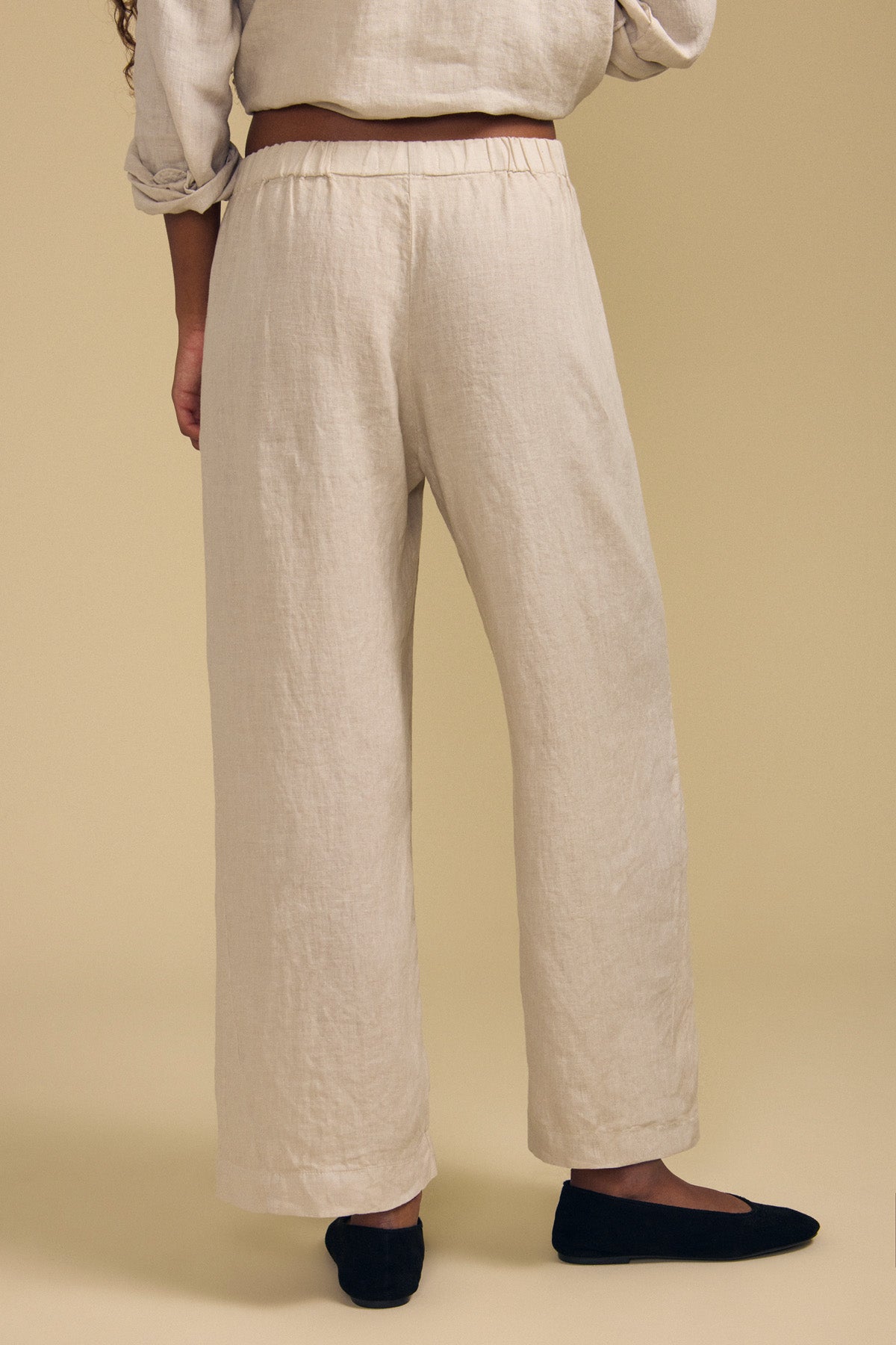A person wearing the LOLA LINEN PANT by Velvet by Graham & Spencer in a loose, light beige wide-leg style with a matching top and black flats, standing against a plain beige background. Only the lower half of the body is visible.-41959882588353