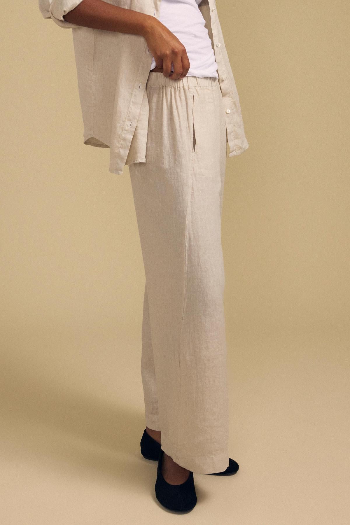 A person dressed in the LOLA LINEN PANT by Velvet by Graham & Spencer—light beige linen pants featuring a relaxed fit and wide leg—paired with a matching open shirt, a white shirt underneath, and black slippers, stands against a plain tan background.