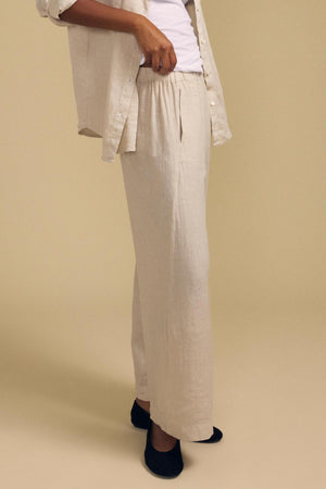 A person dressed in the LOLA LINEN PANT by Velvet by Graham & Spencer—light beige linen pants featuring a relaxed fit and wide leg—paired with a matching open shirt, a white shirt underneath, and black slippers, stands against a plain tan background.