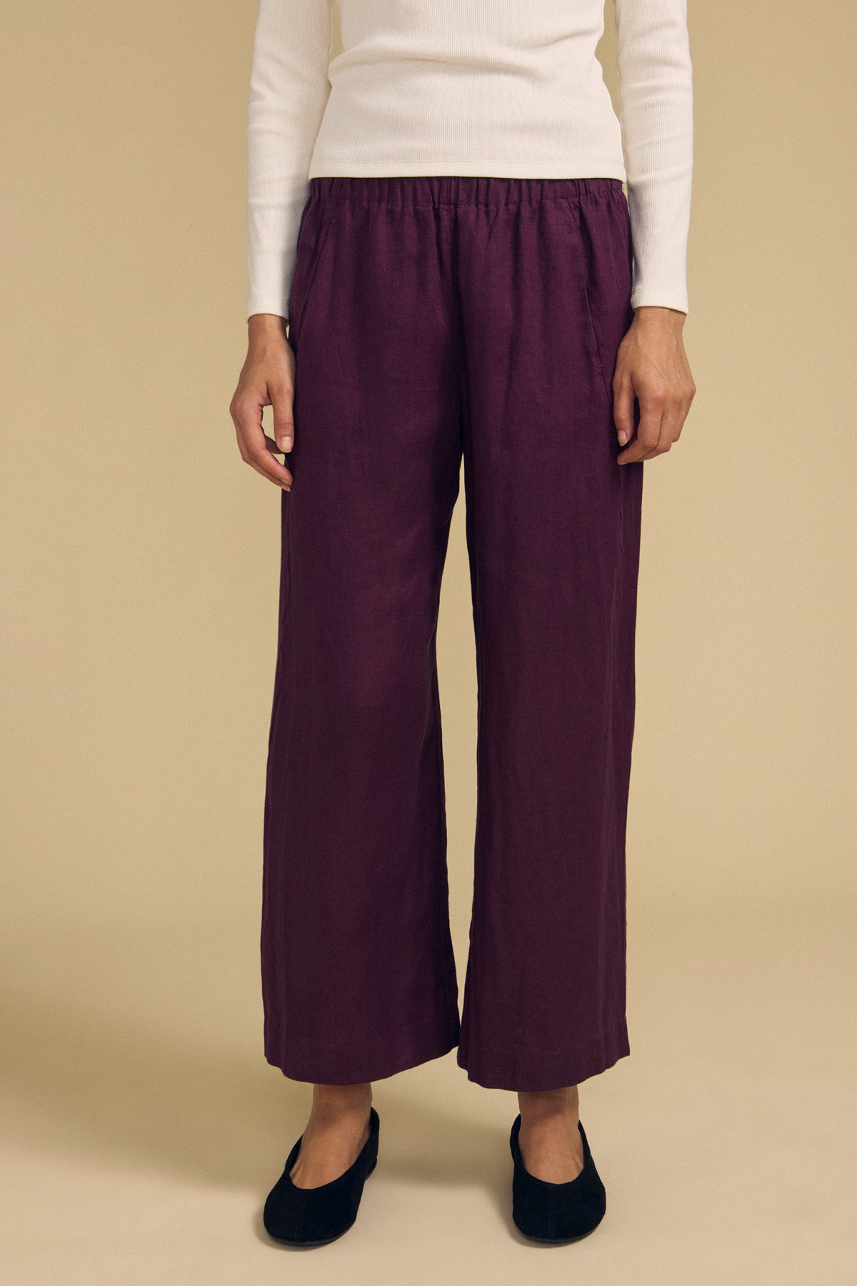 A person stands against a beige background wearing a white long-sleeve top, the Velvet by Graham & Spencer LOLA LINEN PANT in purple—with an elastic waist and wide leg—and black flat shoes.-42063800303809