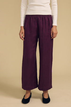 A person stands against a beige background wearing a white long-sleeve top, the Velvet by Graham & Spencer LOLA LINEN PANT in purple—with an elastic waist and wide leg—and black flat shoes.