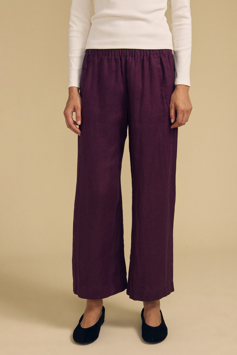 A person stands against a beige background wearing a white long-sleeve top, the Velvet by Graham & Spencer LOLA LINEN PANT in purple—with an elastic waist and wide leg—and black flat shoes.