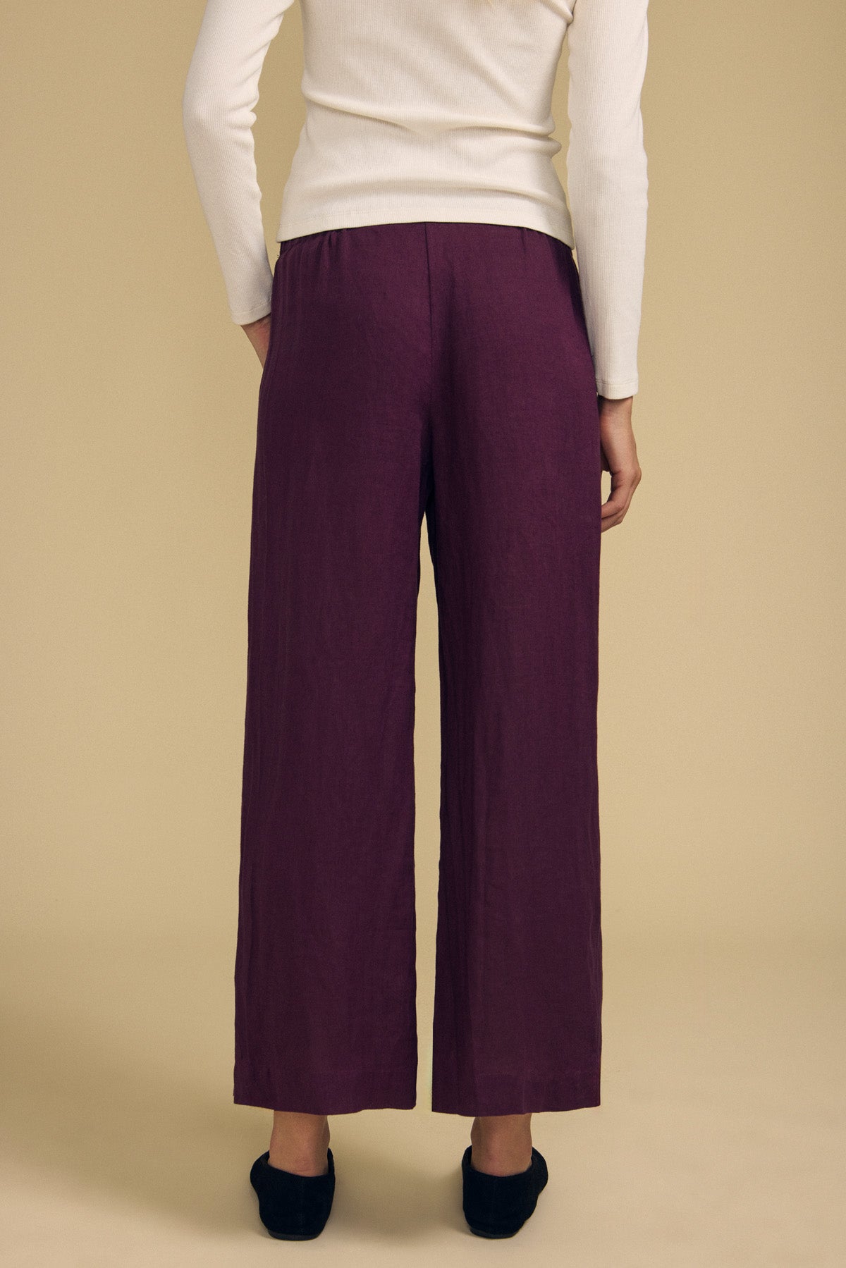 A person stands with their back to the camera against a plain beige background, wearing a white long-sleeve top, black shoes, and the LOLA LINEN PANT in purple by Velvet by Graham & Spencer, featuring a wide-leg silhouette and an elastic waist.