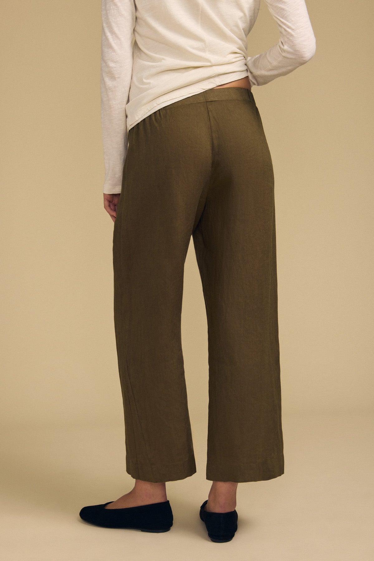 Dressed in the LOLA LINEN PANT by Velvet by Graham & Spencer—an olive-green, wide-leg linen trouser with an elastic waist—paired with a long-sleeve beige top and black slip-on shoes, this person is shown from the back against a plain beige background.