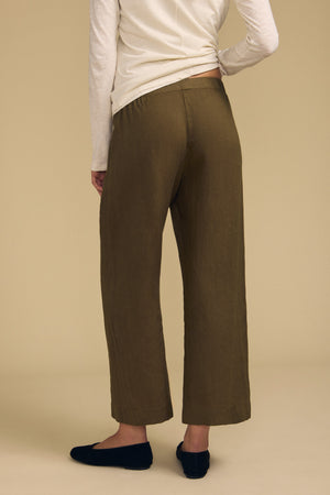 Dressed in the LOLA LINEN PANT by Velvet by Graham & Spencer—an olive-green, wide-leg linen trouser with an elastic waist—paired with a long-sleeve beige top and black slip-on shoes, this person is shown from the back against a plain beige background.