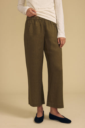 A person wearing an ivory long-sleeve shirt, Velvet by Graham & Spencer’s LOLA LINEN PANT in olive green—featuring a relaxed fit, wide leg silhouette, and elastic waist—and navy flats stands against a beige background.