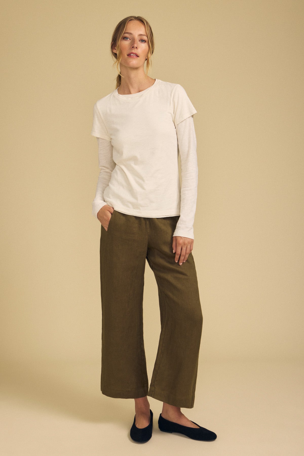 A person stands against a beige background wearing a white layered top, the LOLA LINEN PANT by Velvet by Graham & Spencer—olive-green, wide-leg linen pants with an elastic waist—and black flat shoes, with one hand in their pocket.