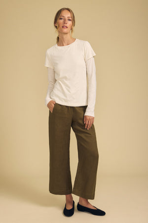 A person stands against a beige background wearing a white layered top, the LOLA LINEN PANT by Velvet by Graham & Spencer—olive-green, wide-leg linen pants with an elastic waist—and black flat shoes, with one hand in their pocket.