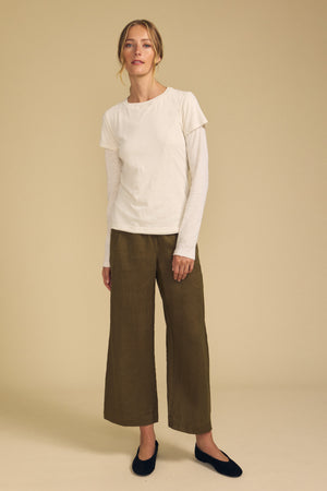 A woman stands against a beige background, wearing a cream long-sleeve top under a short-sleeve t-shirt, paired with Velvet by Graham & Spencer’s LOLA LINEN PANT in olive—a wide-leg linen style with an elastic waist—and black flats.